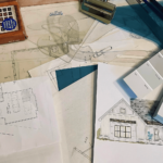 A cluttered desk with architectural drawings, blueprints, a photo of a building exterior, color swatches, and drawing tools scattered across the surface—a true reflection of an interior designer's creative process.